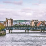 Image of a river with a bridge running across it with buildings and hills in the background