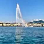 Image of a lake with a large fountain and buildings and mountains in the background