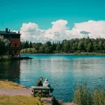 Two men sat by a lake with trees and a blue sky in the background