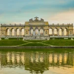 Yellow concrete palace sitting on a small hill behind a body of water