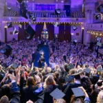 Lots of students in a hall wearing graduation robes throwing their mortarboards in the air to celebrate