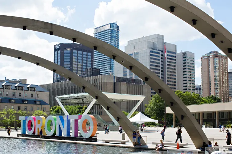 Tall buildings in the background with a white and colourful sign that reads 'Toronto' in all capital letters in the foreground
