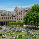 Image of buildings and green trees with a patch of grass with people sat on it