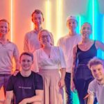 Image of 7 students standing in front of rainbow neon lights smiling at the camera