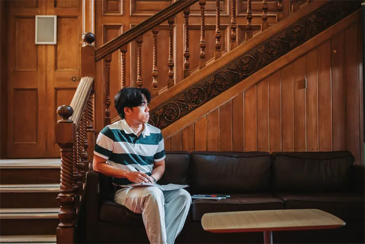 A student sat on a brown sofa at the foot of intricately decorated wooden stairs. They are looking away from the camera and into the distance.