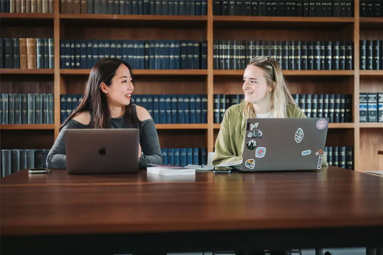 Students sat at a table with their laptops in front of them and a case of books behind them. They are looking at each other and smiling.