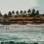 A beach with palm trees and shack style buildings on the shore, with the sea lapping up against the sand