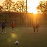Image of 6 footballers standing in a line on a football pitch, with footballs in the foreground and a sunset in the background