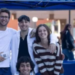 Three students stood next to each other, with one student sitting in front of them, all smiling at the camera