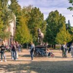 Image of people walking around in a courtyard with green trees and a horse statue