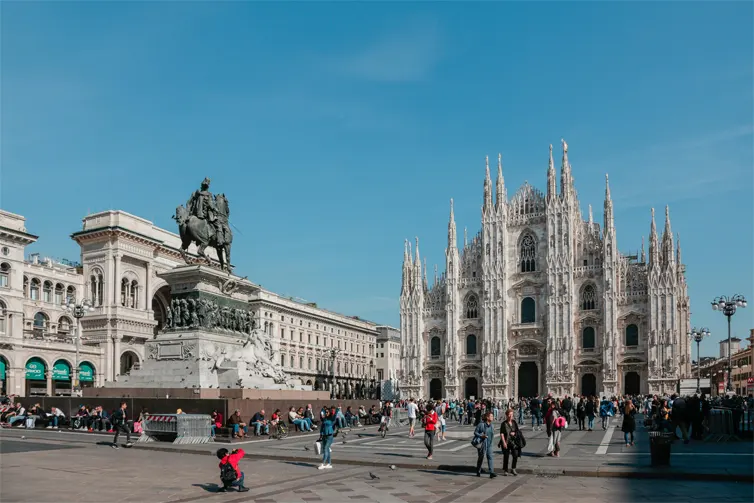 Large statue and cathedral on a city square with people standing around and taking photographs.