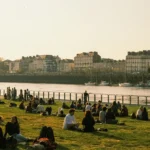 Image of people sat on grass next to a river, with a row of buildings the other side of the river.