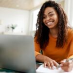 Image of a student wearing a bright orange jumper smiling at a laptop screen