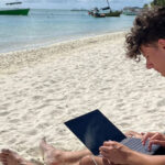 A student sat on a white sandy beach looking at their laptop which is resting on their lap