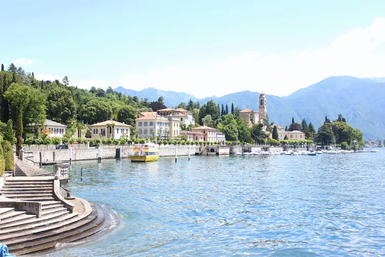The shore of a lake, with buildings and trees in the foreground and mountains in the background.