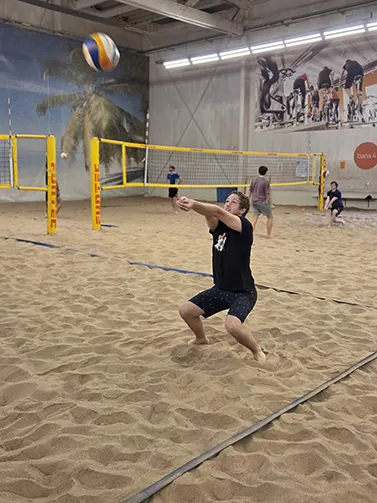 Image of a student wearing a black t-shirt and shorts, playing beach volleyball in a sandy sports hall