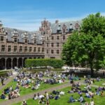 Image of a large, intricately designed building with a grass courtyard in front, with lots of people sat down in groups on the grass