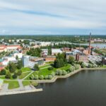 Image of several redbrick and glass buildings with greenery around them. There is a body of water in the foreground and a city landscape in the background